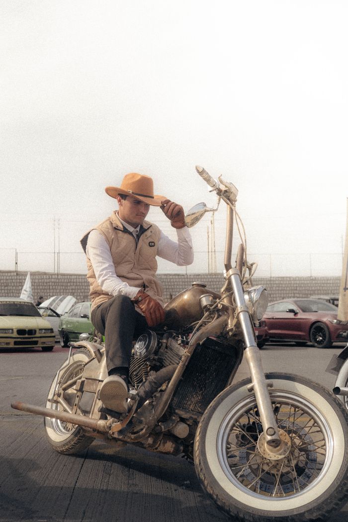 Man in cowboy hat sitting on a motorcycle in an outdoor parking lot during the day.