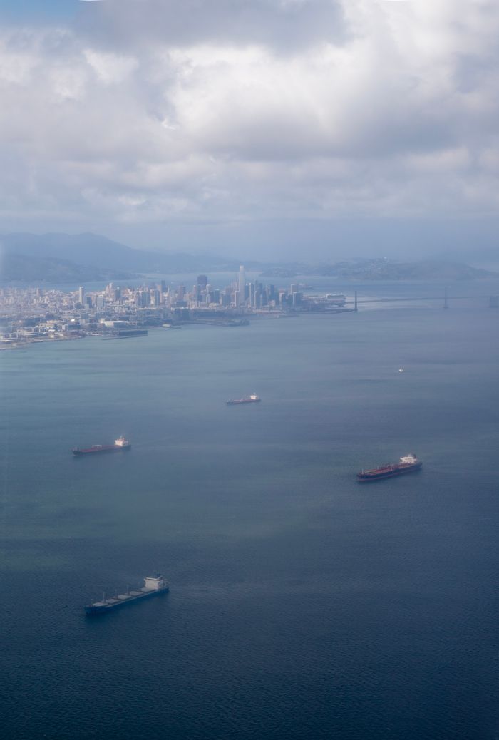 about-img-01 Aerial shot of cargo ships in San Francisco Bay with city skyline and iconic bridge in the background.
