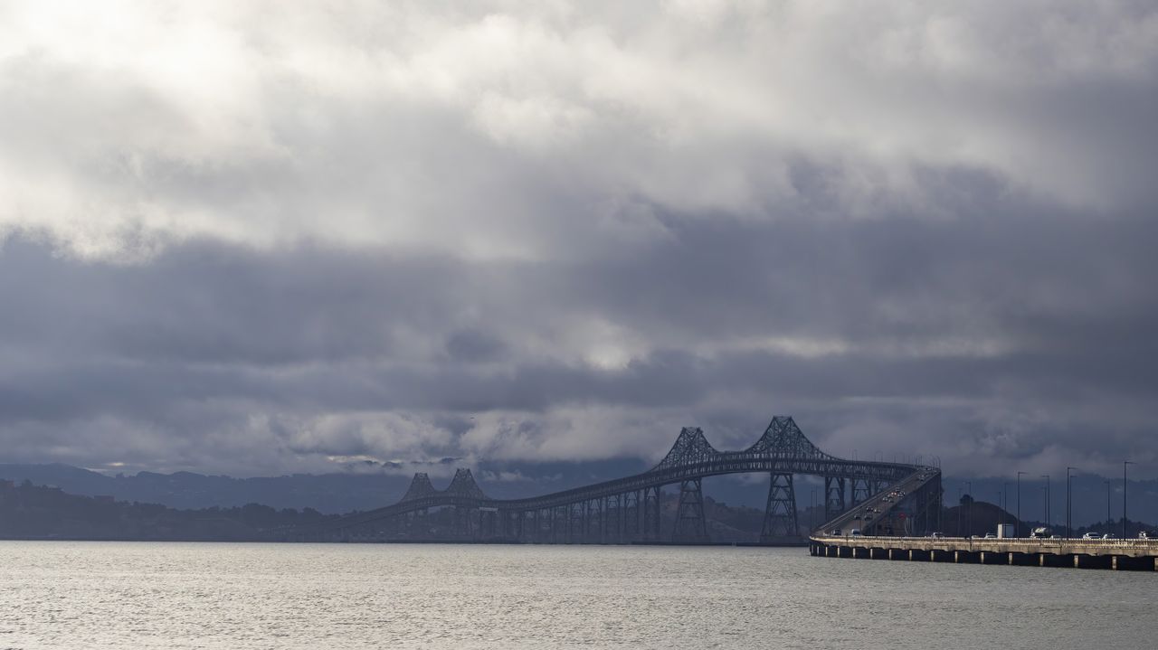 services-01 Aerial view of a bridge under an overcast sky, reflecting its silhouette on the calm water.